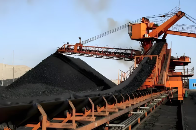 A large industrial machine transporting coal, with a huge pile of coal in the background and a clear blue sky.